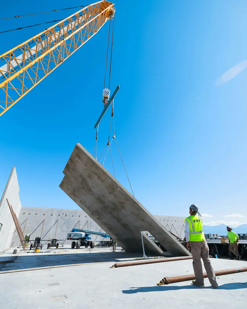 Ein Kran hebt ein Betonfertigteil, ein Arbeiter in Warnweste richtet es aus